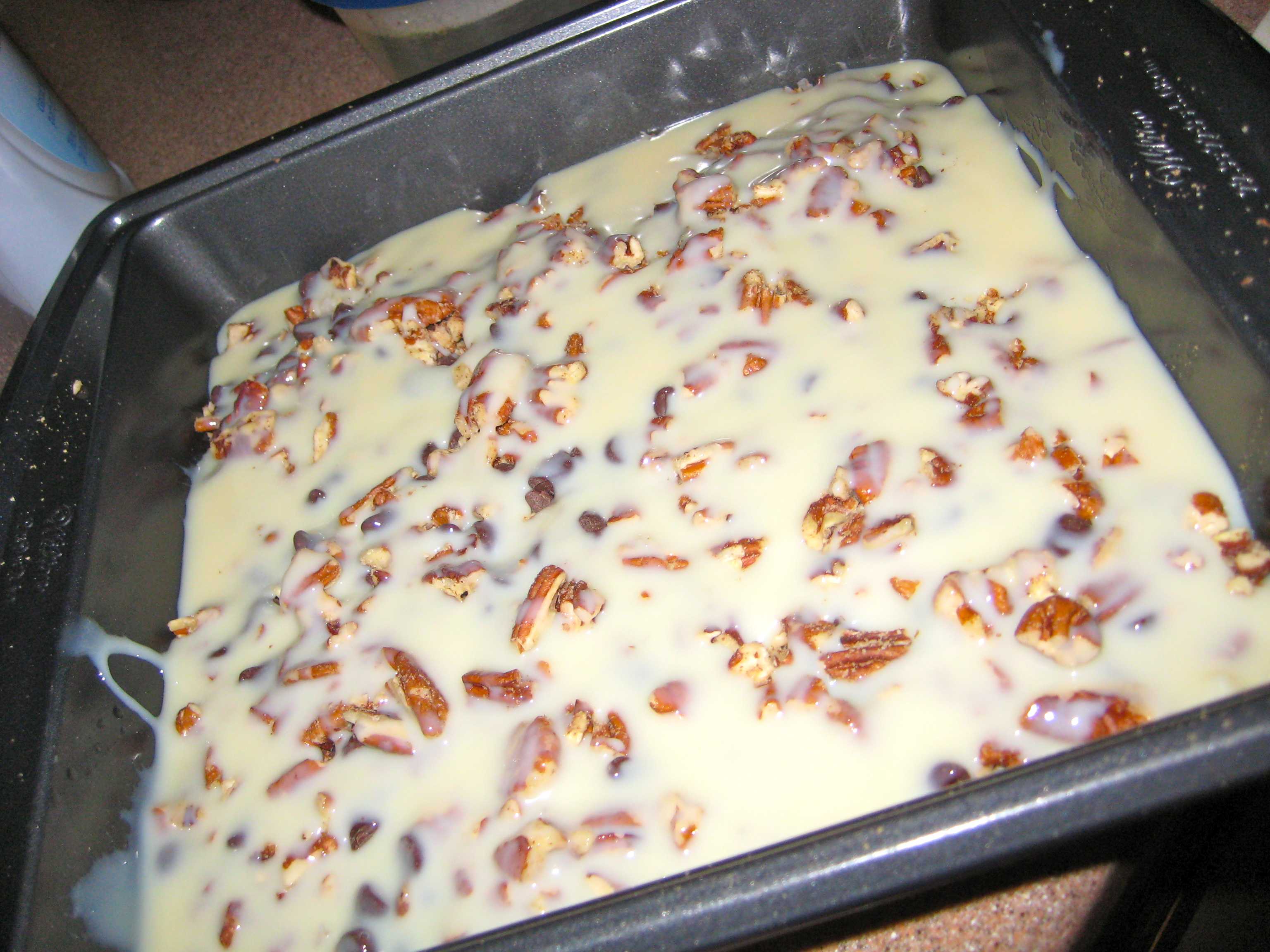 Overhead view of a baking pan with condensed milk poured over pecans and chocolate chips, warm golden tones evoking a cozy breakfast kitchen