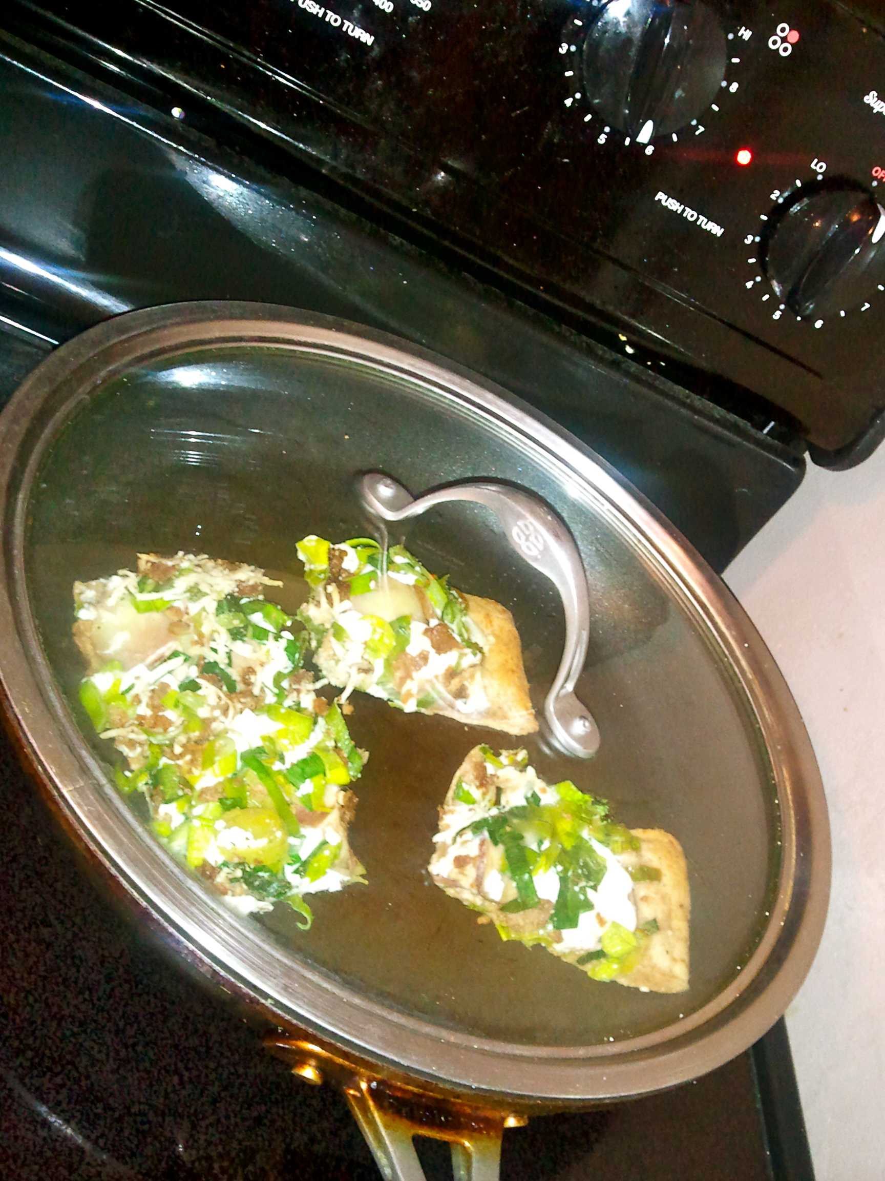 Overhead view of a pan with a glass lid containing pizza topped flatbread slices with green onions and cheese, cozy kitchen scene