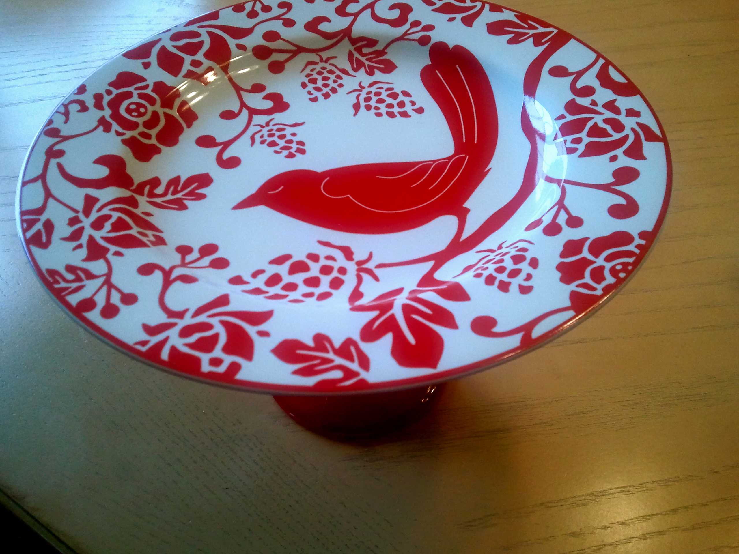 Decorative red and white pedestal plate with a bird and floral pattern resting on a wooden table, warm nostalgic mood