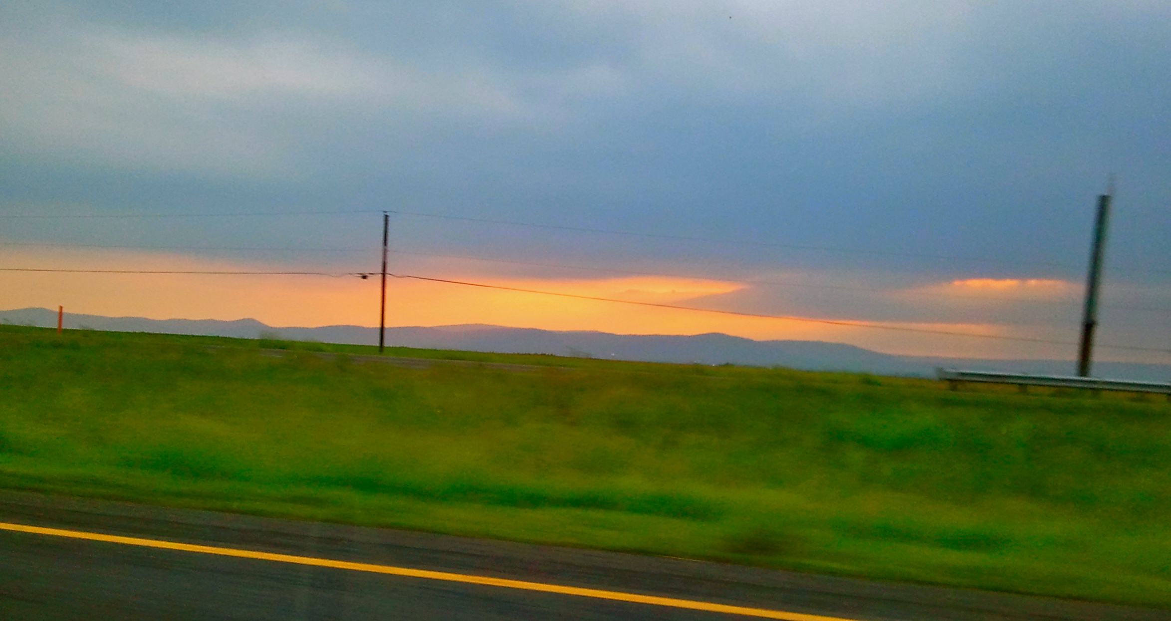 A blurred roadside view of green fields and distant hills under a dramatic sunset sky with orange and blue tones